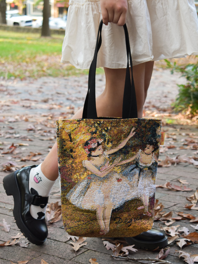 Person holding a tote bag with a Degas Two Dancers painting design on a leafy street.