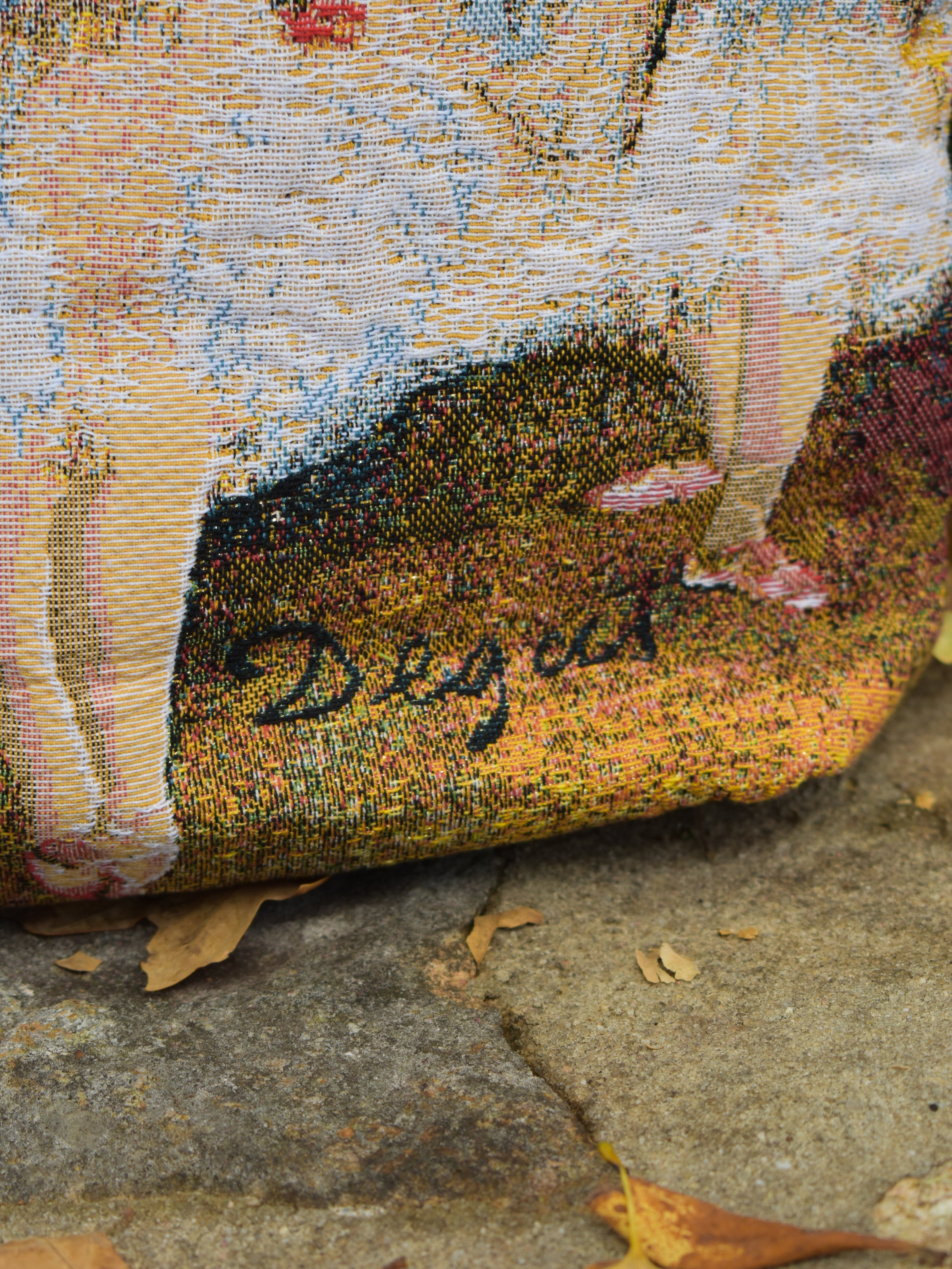 Close-up of a textured fabric bag with a Degas signature on a stone background