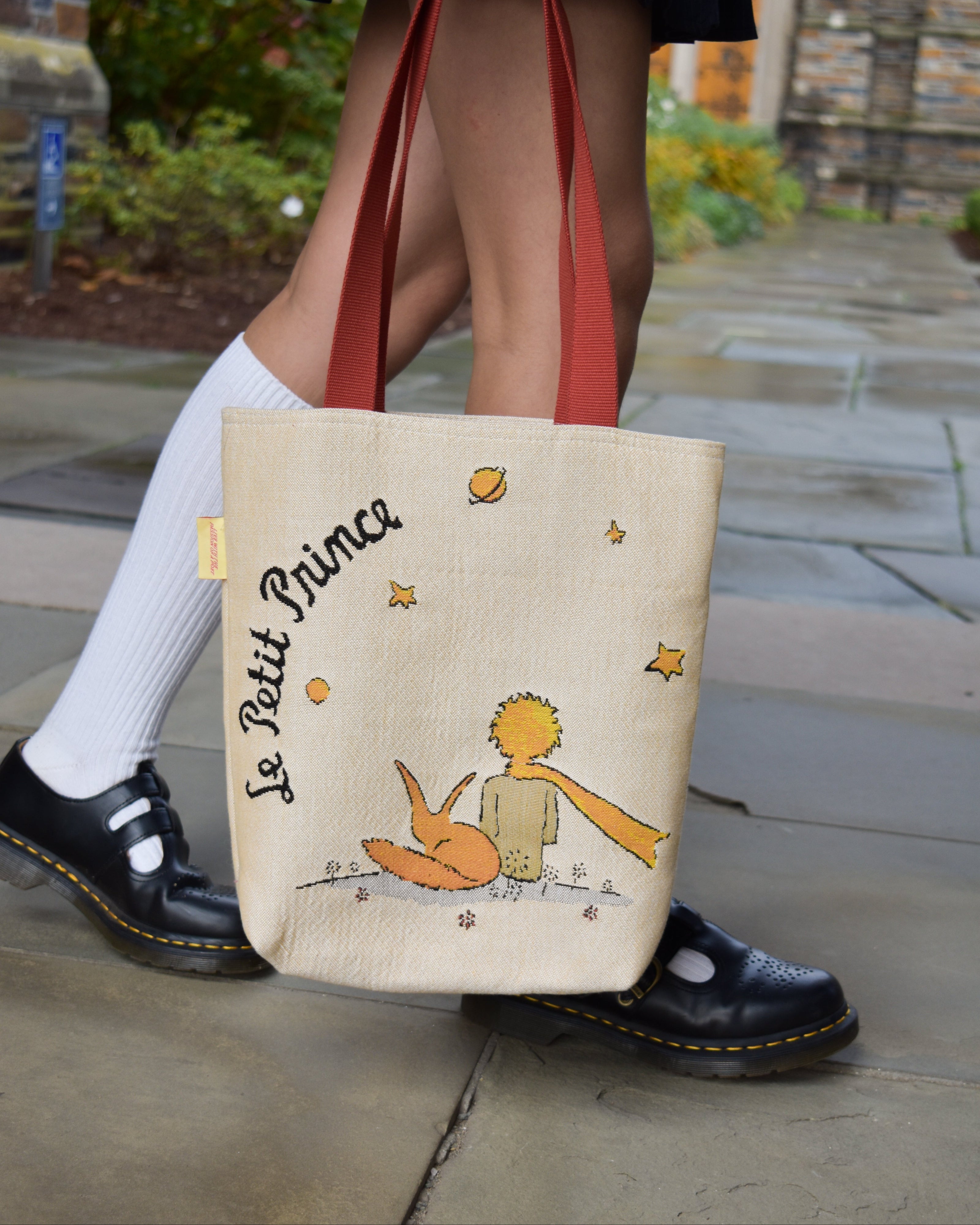 Tote bag with 'Le Petit Prince' design held by a person wearing white socks and black shoes.