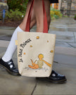 Tote bag with 'Le Petit Prince' design held by a person wearing white socks and black shoes.