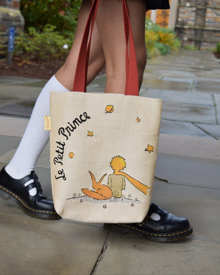 Tote bag with 'Le Petit Prince' design held by a person wearing white socks and black shoes.