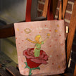 Tote bag with embroidered design of a child and rose on a wooden chair.
