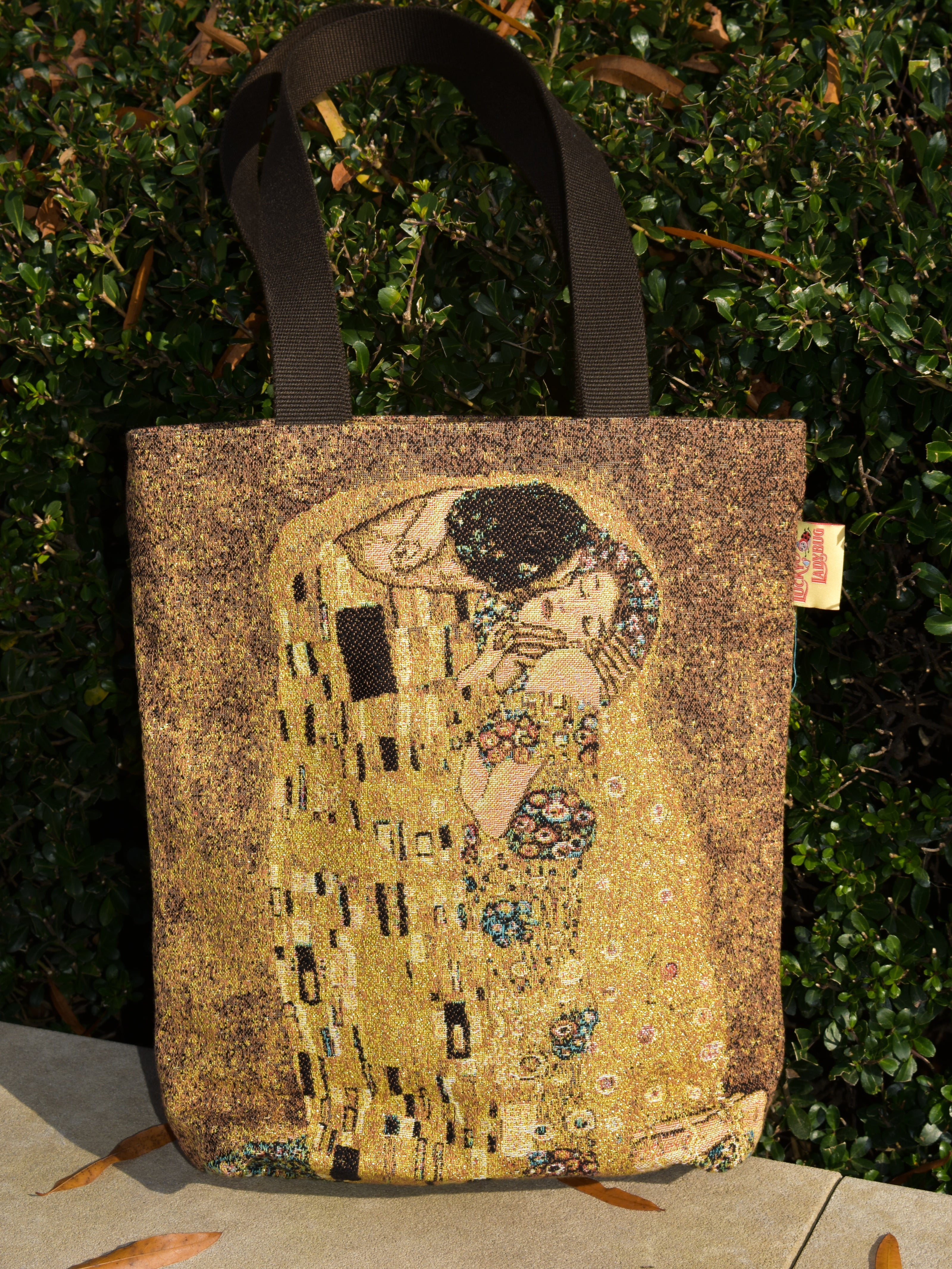 Tote bag with 'The Kiss' artwork by Gustav Klimt against a green bush background